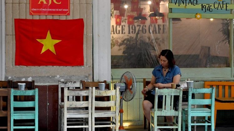 Eine Frau schaut aufs Handy während sie in Hanoi einen Kaffee trinkt, am Laden ist eine vietnamesische Flagge angebracht