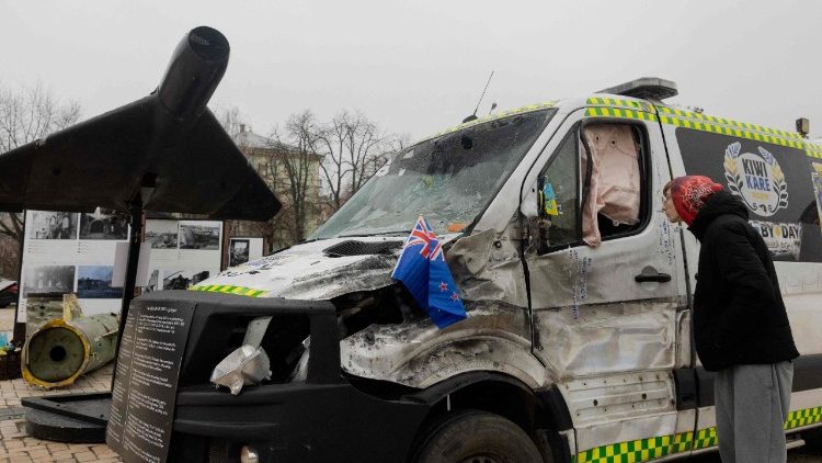 Um homem observa uma ambul&acirc;ncia Kiwi K.A.R.E. destru&iacute;da, parte do projeto humanit&aacute;rio "A Estrada da Vida" entre a Nova Zel&acirc;ndia e a Ucr&acirc;nia, atingida por um drone russo durante uma miss&atilde;o de evacua&ccedil;&atilde;o em junho de 2025. A ambul&acirc;ncia est&aacute; ao lado de um drone Shahed 136 (Geranium-2) de fabrica&ccedil;&atilde;o iraniana, pertencente ao Ex&eacute;rcito Russo, em uma exposi&ccedil;&atilde;o a c&eacute;u aberto de equipamentos militares russos destru&iacute;dos na Pra&ccedil;a Mykhailivska (Pra&ccedil;a de S&atilde;o Miguel), em Kiev, em 9 de dezembro de 2025, em meio &agrave; invas&atilde;o russa da Ucr&acirc;nia. (Foto de Tetiana DZHAFAROVA / AFP)