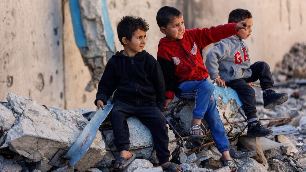 Crianças palestinas deslocadas que vivem nas proximidades da sede da Agência das Nações Unidas de Assistência aos Refugiados da Palestina no Oriente Próximo (UNRWA) sentam-se sobre os escombros de um prédio destruído, na Cidade de Gaza, em 11 de janeiro de 2026. (Foto de Omar AL-QATTAA / AFP)