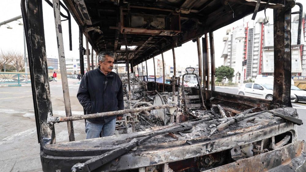 Um homem está ao lado dos destroços de um ônibus incendiado na Praça Sadeghieh, em Teerã, em 15 de janeiro de 2026. Um movimento de protesto em todo o Irã, inicialmente desencadeado por queixas econômicas, transformou-se em um dos maiores desafios à liderança clerical desde que assumiu o poder em 1979. (Foto de ATTA KENARE / AFP)