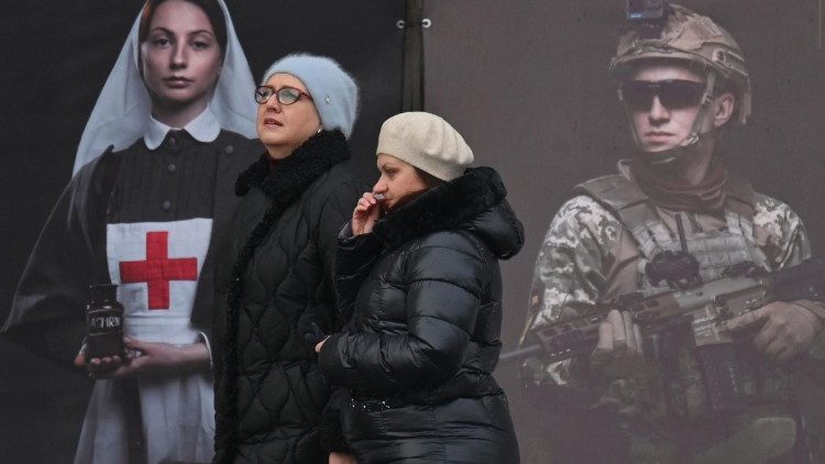 Mulheres caminham em frente a um cartaz exibido pelo Instituto Nacional da Mem&oacute;ria Nacional como parte de uma exposi&ccedil;&atilde;o ao ar livre no centro de Kiev, em 8 de fevereiro de 2026, em meio &agrave; invas&atilde;o russa na Ucr&acirc;nia. (Foto de Sergei SUPINSKY / AFP)