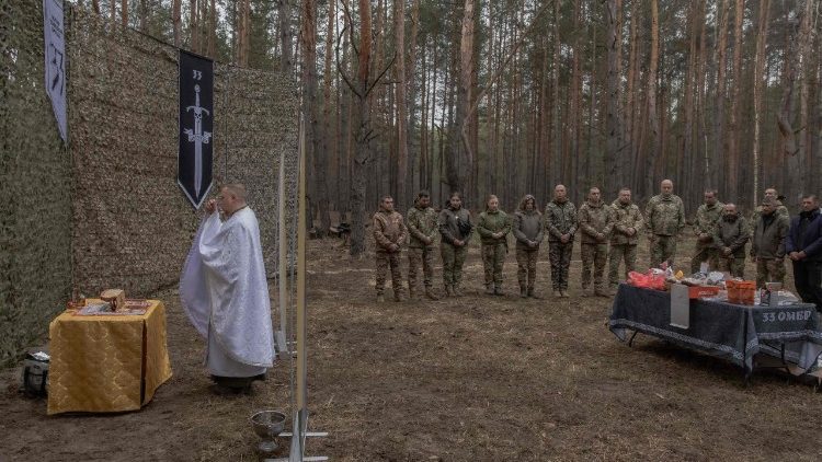 O capel&atilde;o militar Mykola Bahirov reza com membros do 33&ordm; Batalh&atilde;o Mecanizado Independente da Ucr&acirc;nia, ao lado de bolos e outros alimentos tradicionais, para celebrar a P&aacute;scoa Ortodoxa, na regi&atilde;o de Kharkiv, em 12 de abril de 2026, em meio &agrave; invas&atilde;o russa da Ucr&acirc;nia. Greco-cat&oacute;lico, este homem de 39 anos trabalhava como secret&aacute;rio diocesano quando a R&uacute;ssia lan&ccedil;ou sua invas&atilde;o em grande escala da Ucr&acirc;nia, em fevereiro de 2022. Foi no funeral de um soldado, ao presenciar o terror nas filhas do falecido, que Mykola Bahirov decidiu se tornar capel&atilde;o: em vez de acompanh&aacute;-las na morte, ele permaneceria, enquanto elas ainda estivessem vivas, perto dos soldados ucranianos que lutavam contra o invasor russo. (Foto de Roman PILIPEY / AFP)