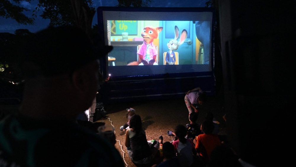 Pessoas deslocadas, incluindo crianças, assistem a um filme de animação projetado em uma tela ao ar livre em Caraballeda, estado de La Guaira, Venezuela, em 4 de julho de 2026, após os terremotos gêmeos. Os devastadores terremotos gêmeos na Venezuela mataram quase 3.000 pessoas, segundo dados oficiais divulgados em 4 de julho, enquanto equipes internacionais de resgate começavam a encerrar as operações de busca por sobreviventes nos escombros. (Foto de Juan Barreto / AFP)