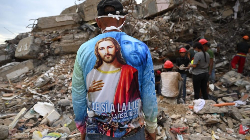 Uma pessoa vestindo uma camisa com a imagem de Jesus está em frente a equipes de resgate que vasculham os escombros de um prédio de apartamentos que desabou em Caraballeda, estado de La Guaira, Venezuela, em 12 de julho de 2026, após os terremotos gêmeos que atingiram a região. Os terremotos que atingiram a Venezuela em 24 de junho deixaram quase 4.500 mortos até 12 de julho, enquanto grande parte da população afetada continuava vivendo em acampamentos, segundo dados oficiais. (Foto de Raul Arboleda / AFP)
