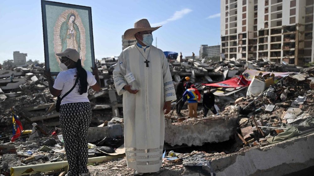 Fiel venezuelana ao lado de sacerdote segura uma imagem da Virgem de Guadalupe enquanto voluntários e familiares das vítimas procuram pelos corpos de seus entes queridos sob os escombros de um prédio destruído pelos terremotos de 24 de junho em Caraballeda, Venezuela, em 17 de julho de 2026. (Foto de Juan Barreto / AFP)