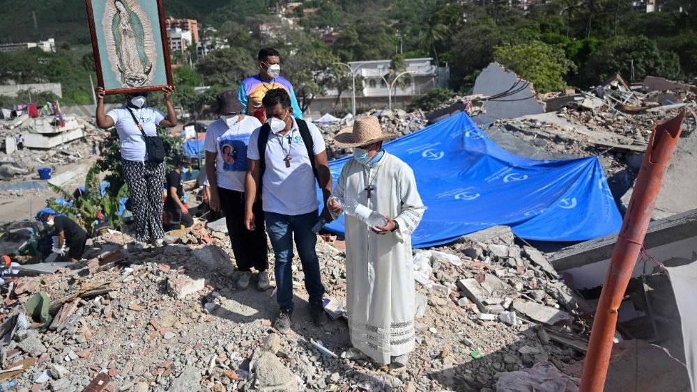 Um padre asperge água benta sobre os escombros de um prédio desabado enquanto uma mulher segura uma imagem da Virgem de Guadalupe e outros fiéis observam de perto em Caraballeda, Venezuela, em 17 de julho de 2026, após os terremotos de 24 de junho. (Foto de Juan Barreto / AFP)