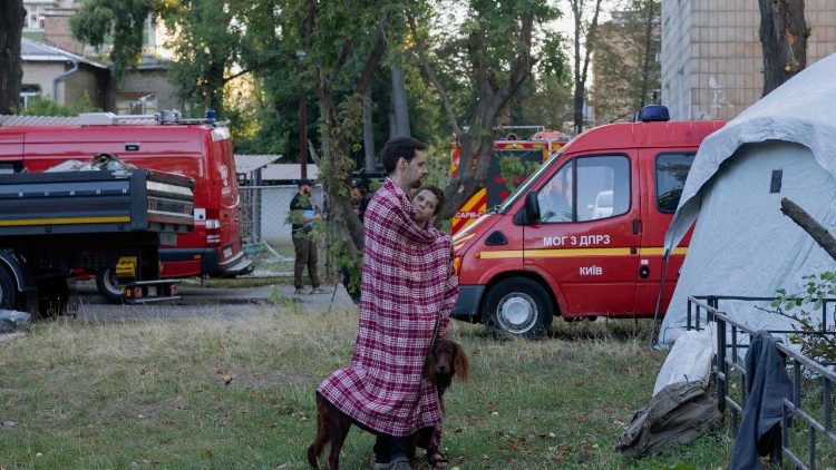 Um homem est&aacute; com um menino e um cachorro debaixo de um cobertor perto de um pr&eacute;dio residencial danificado ap&oacute;s um ataque russo em Kiev, em 20 de agosto de 2026, em meio &agrave; invas&atilde;o russa da Ucr&acirc;nia. O bombardeio russo matou seis pessoas e feriu dezenas na capital ucraniana e na regi&atilde;o circundante durante a noite, disseram as autoridades em 20 de agosto. (Foto de Tetiana DZHAFAROVA / AFP)  