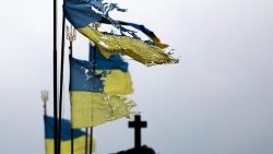 Bandeiras ucranianas rasgadas agitadas pelo vento, tendo ao fundo a cruz de uma igreja.