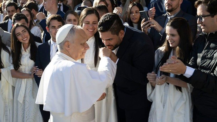 Matheus de Macêdo beija o anel de pescador do Santo Padre, enquanto Amanda de Macêdo, seu lado direito, olha e sorri. 